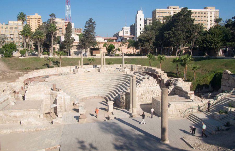 The ruins of the Roman Amphitheatre Alexandria Egypt, showing the white marble seating tiers of the ancient structure, surrounded by green grass and modern city buildings. This stop is key for any Egypt travel itinerary 10 days.