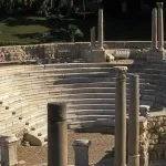 The ancient stone seating tiers and ruins of the Roman Amphitheatre Alexandria, Egypt, showing partially restored columns and surrounding green gardens under natural light.
