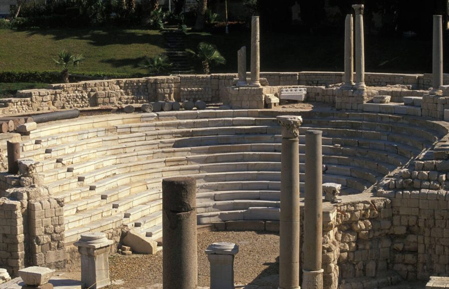 The ancient stone seating tiers and ruins of the Roman Amphitheatre Alexandria, Egypt, showing partially restored columns and surrounding green gardens under natural light.