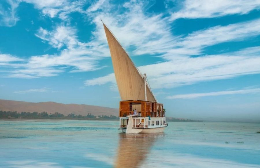 A traditional Egyptian two-masted sailing boat, a dahabiya, sailing on the Nile River under blue skies.