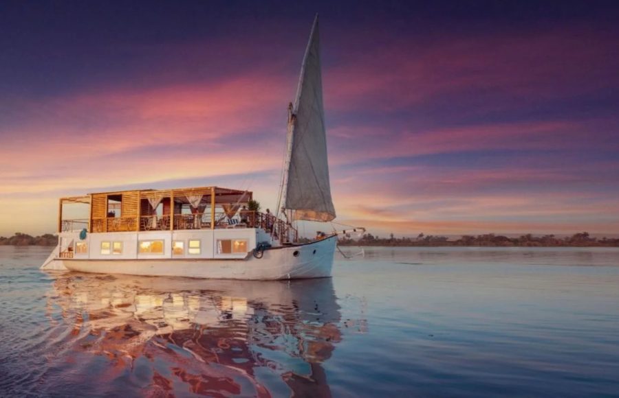 traditional two-masted Dahabiya sailboat with people on board cruises the Nile River at sunset.