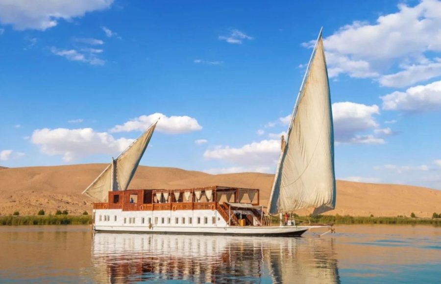 A traditional two-masted Dahabiya sailboat cruises the Nile River past a sandy riverbank.