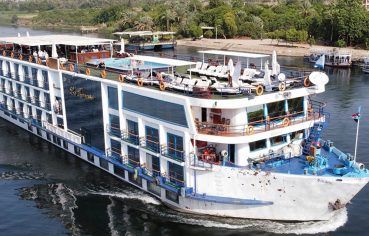 A large white Nile cruise ship on the water near a lush green bank.