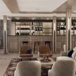 Modern cruise ship bar and lounge area with light wood paneling, a bar counter, and various seating groups with velvet upholstery.