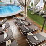 Overhead view of a cruise sundeck with a blue circular pool, wooden decking, artificial turf, and rows of modern gray lounge chairs.