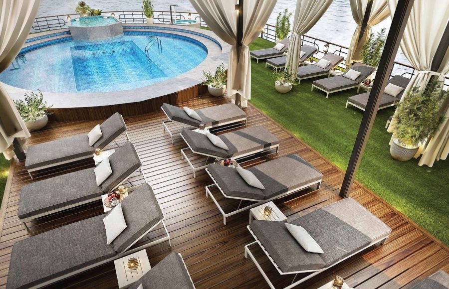 Overhead view of a cruise sundeck with a blue circular pool, wooden decking, artificial turf, and rows of modern gray lounge chairs.