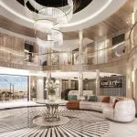 Wide view of a luxurious, circular, two-story cruise ship atrium with curved sofas, a grand chandelier, and light wood and gold accents.