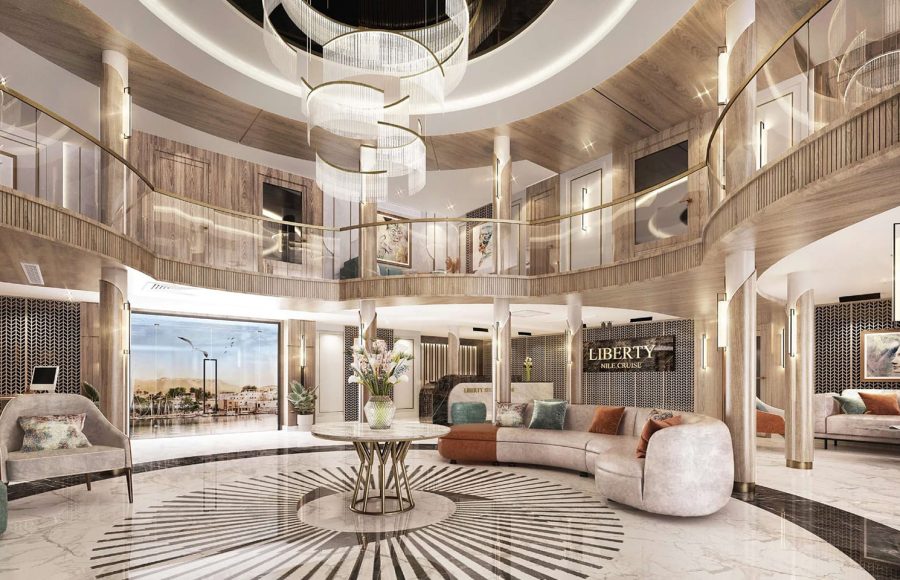 Wide view of a luxurious, circular, two-story cruise ship atrium with curved sofas, a grand chandelier, and light wood and gold accents.