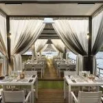Covered outdoor dining area on a cruise deck with white tables, sheer curtains, and a sailboat visible on the water.