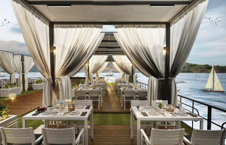 Covered outdoor dining area on a cruise deck with white tables, sheer curtains, and a sailboat visible on the water.