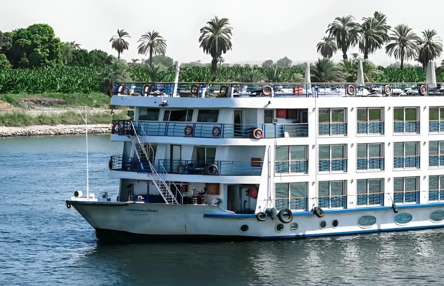 A white multi-deck cruise ship sailing on the River Nile with palm trees on the bank.