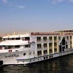 The M/S Royal Viking Nile cruise ship docked alongside a stone quay, showing its multi-deck white and tan exterior, with Arabic text on the bow and passengers visible on the upper decks.