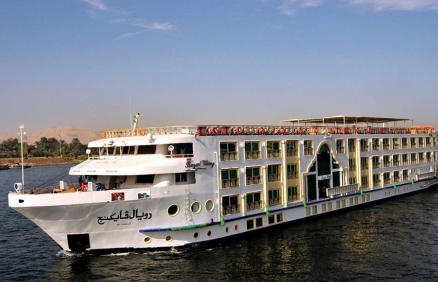 The M/S Royal Viking Nile cruise ship docked alongside a stone quay, showing its multi-deck white and tan exterior, with Arabic text on the bow and passengers visible on the upper decks.