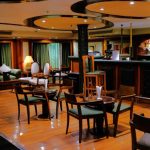 The interior of the Royal Viking Nile Cruise main lounge, featuring a wooden bar, small round tables with chairs, and a seating area with green sofas.