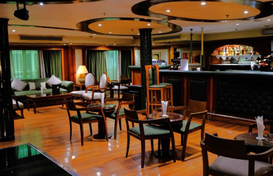 The interior of the Royal Viking Nile Cruise main lounge, featuring a wooden bar, small round tables with chairs, and a seating area with green sofas.