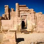 The sun-drenched, partially reconstructed mud-brick and stone facade of the Ruins Of Deir El Haggar Temple At Kharga Oasis, showing the central doorway and crumbling structures against a bright blue sky.