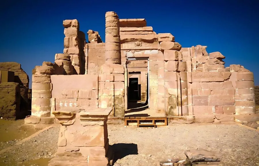 The sun-drenched, partially reconstructed mud-brick and stone facade of the Ruins Of Deir El Haggar Temple At Kharga Oasis, showing the central doorway and crumbling structures against a bright blue sky.