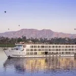 A panoramic view of the Sphinx Nile Cruise ship sailing on the serene Nile River with hot air balloons and mountains in the background