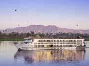 A panoramic view of the Sphinx Nile Cruise ship sailing on the serene Nile River with hot air balloons and mountains in the background