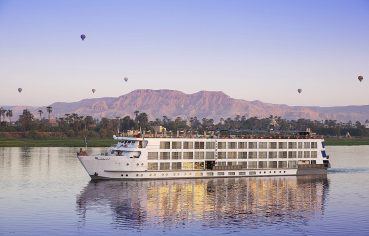 A panoramic view of the Sphinx Nile Cruise ship sailing on the serene Nile River with hot air balloons and mountains in the background