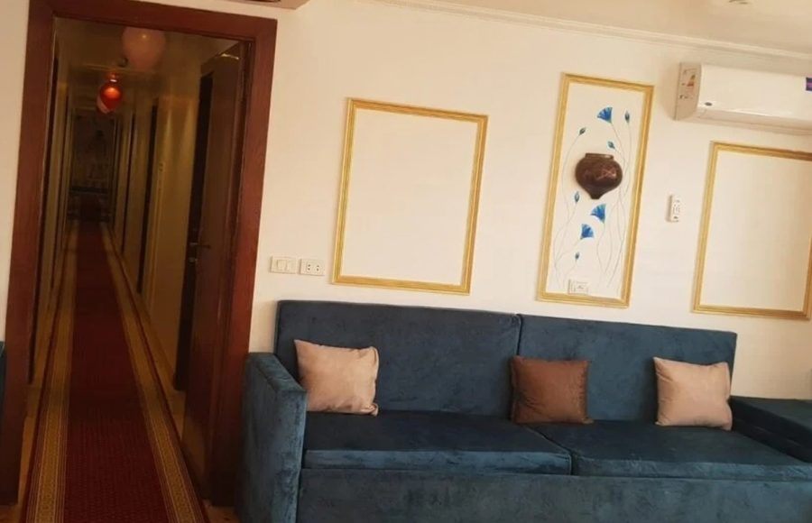 A blue sofa with decorative pillows in a cruise ship cabin, next to a hallway with a red carpet.