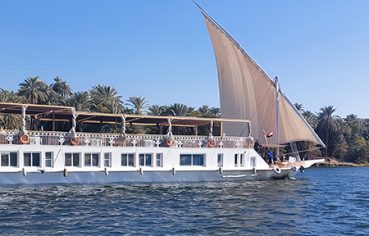 Short Alternative Text: A blue sofa with three throw pillows inside a cabin next to an open door revealing a long hallway with a red runner carpet.