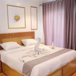 A large bed in a cruise cabin featuring a white towel swan sculpture, wooden headboard, and soft purple curtains.