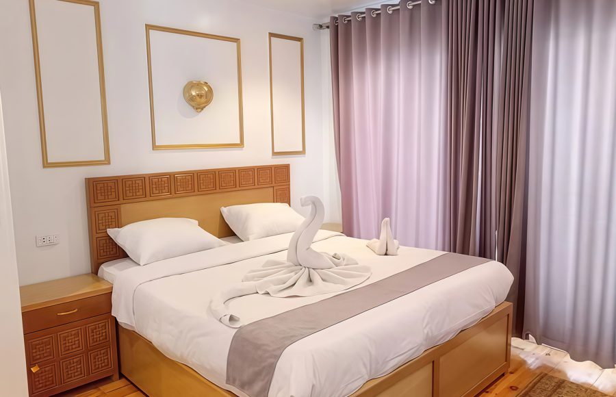 A large bed in a cruise cabin featuring a white towel swan sculpture, wooden headboard, and soft purple curtains.