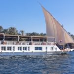Short Alternative Text: A blue sofa with three throw pillows inside a cabin next to an open door revealing a long hallway with a red runner carpet.