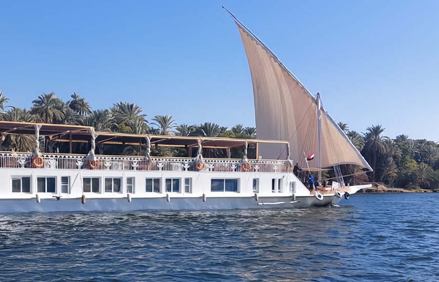 Short Alternative Text: A blue sofa with three throw pillows inside a cabin next to an open door revealing a long hallway with a red runner carpet.