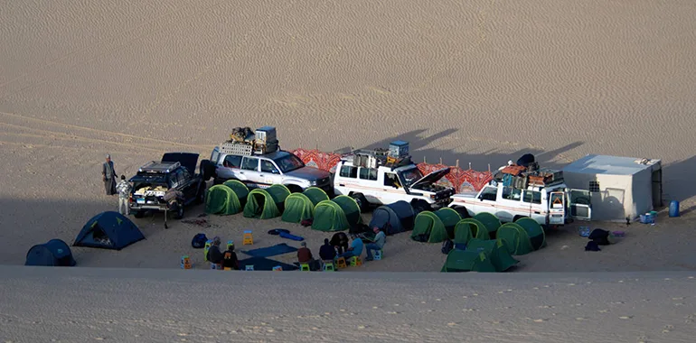 A Line Of Four 4X4 Safari Vehicles Traveling Across A Vast, Golden Sand Plain, Symbolizing A Remote Desert And Oasis Adventure.