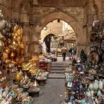 A busy, arched stone alleyway in the Khan El Khalili Bazaar Cairo Egypt, lined with shops displaying rows of colorful lamps, glassware, and metallic artisan goods.