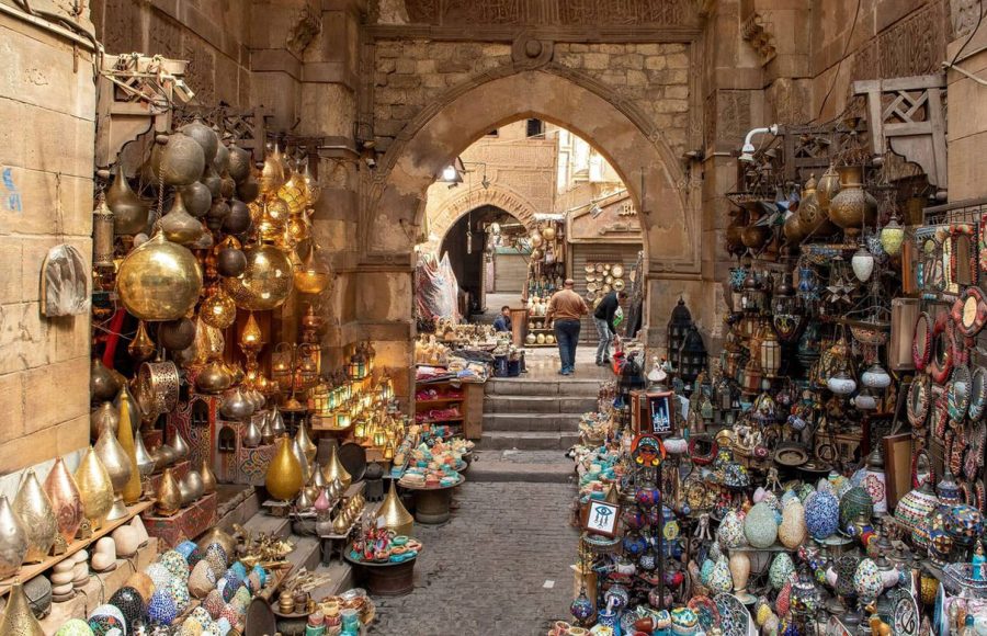 A busy, arched stone alleyway in the Khan El Khalili Bazaar Cairo Egypt, lined with shops displaying rows of colorful lamps, glassware, and metallic artisan goods.
