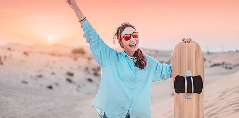 Smiling Female Tourist Is Sandboarding In The Desert At Sunset Holding A Board And Making Victory Sign