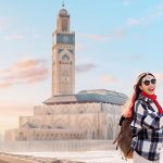 Smiling-tourist-capturing-the-stunning-Hassan-II-Mosque-by-phone-at-sunset-surrounded-by-the-vibrant-atmosphere-of-the-Casablanca-Morocco