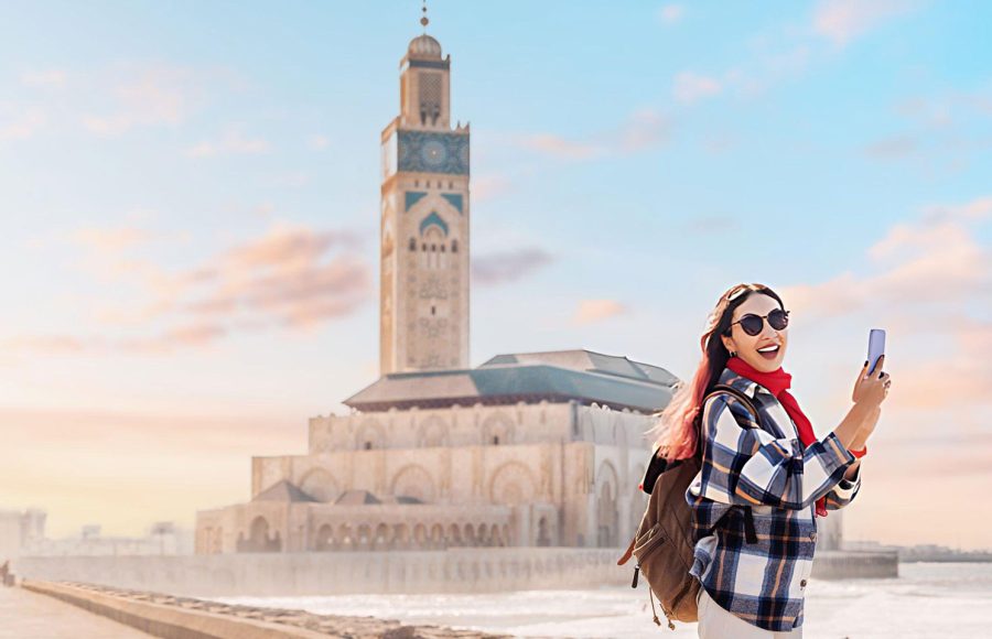 Smiling-tourist-capturing-the-stunning-Hassan-II-Mosque-by-phone-at-sunset-surrounded-by-the-vibrant-atmosphere-of-the-Casablanca-Morocco