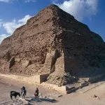 The large, weathered tiers of the Saqqara step pyramid standing in the desert under a clear blue sky, with two people on horseback in the foreground.