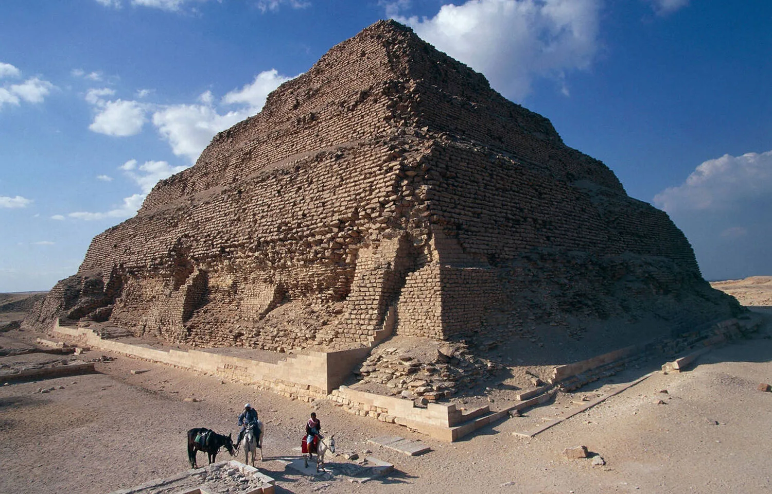 The large, weathered tiers of the Saqqara step pyramid standing in the desert under a clear blue sky, with two people on horseback in the foreground.