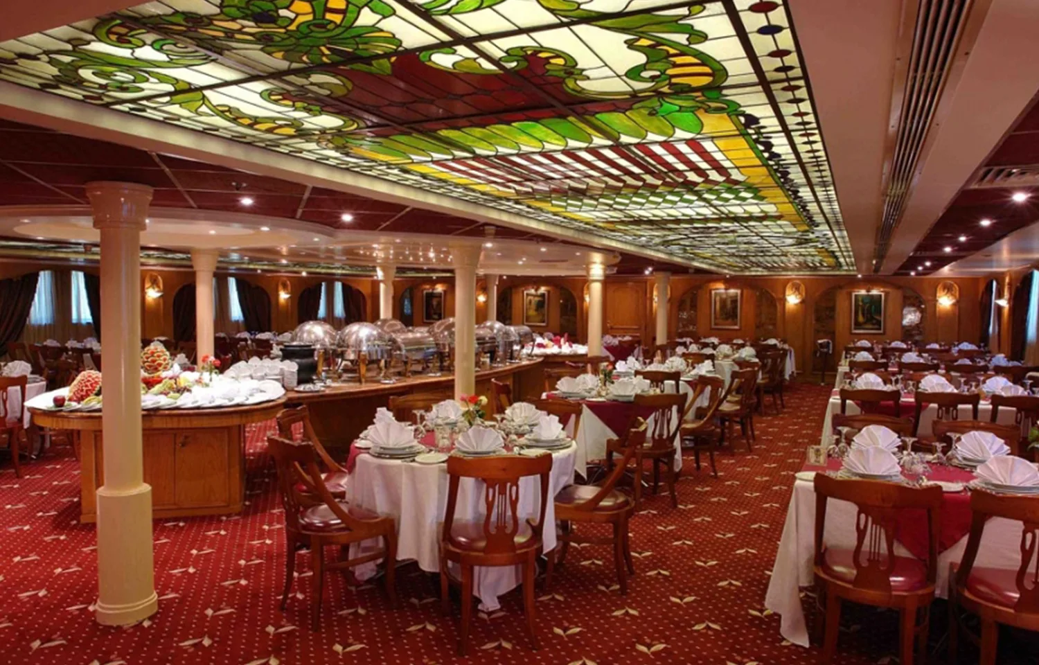 Interior view of a classic dining room on a Nile cruise ship, featuring a large stained-glass ceiling, red patterned carpet, and set round tables.