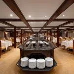 Modern, spacious dining room on a cruise ship with a large central buffet island, dark wood paneling, and a light tan carpet.