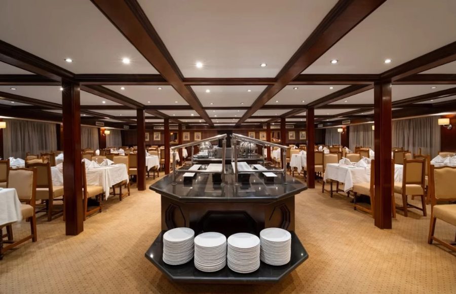 Modern, spacious dining room on a cruise ship with a large central buffet island, dark wood paneling, and a light tan carpet.