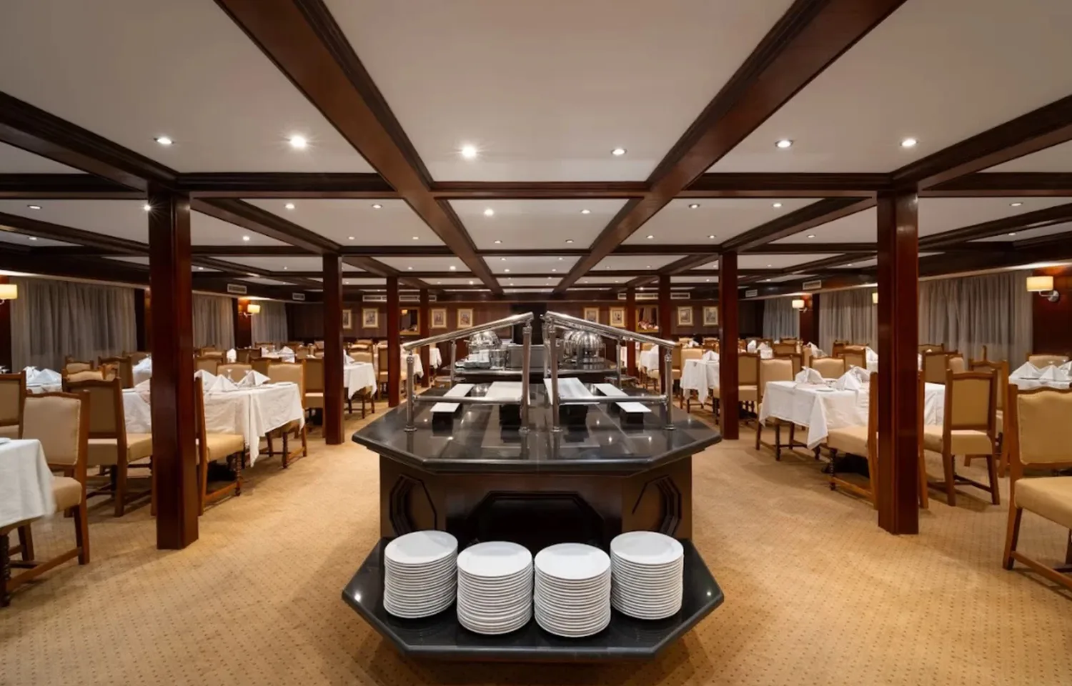 Modern, spacious dining room on a cruise ship with a large central buffet island, dark wood paneling, and a light tan carpet.