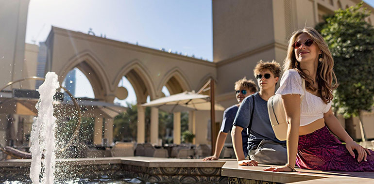 Teenage tourists relaxe on the edge of a fountain amid outdoor cafes in Downtown Dubai