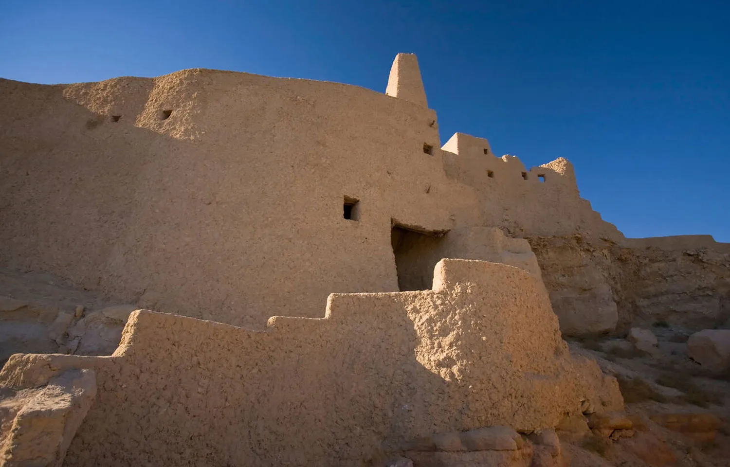 The large, mud-brick ruins of the Tempe of Amun Aghurmi acropolis Siwa Oasis Egypt, featuring a tall, tapered tower-like structure and a stepped base, against a deep blue sky.