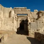 The ancient, light-colored limestone ruins of the Temple of the Oracle Aghurmi in Siwa, Egypt, framed against a deep blue sky.