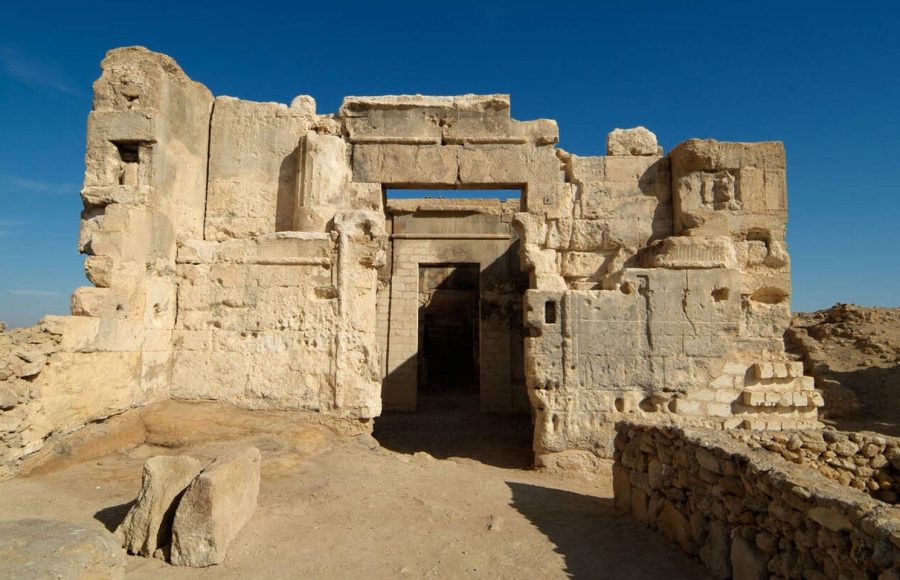 The ancient, light-colored limestone ruins of the Temple of the Oracle Aghurmi in Siwa, Egypt, framed against a deep blue sky.