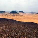 An expansive view of the Black Desert in Egypt, showing numerous conical, volcano-shaped black mounds contrasting with the ochre-colored desert sand under an overcast sky.