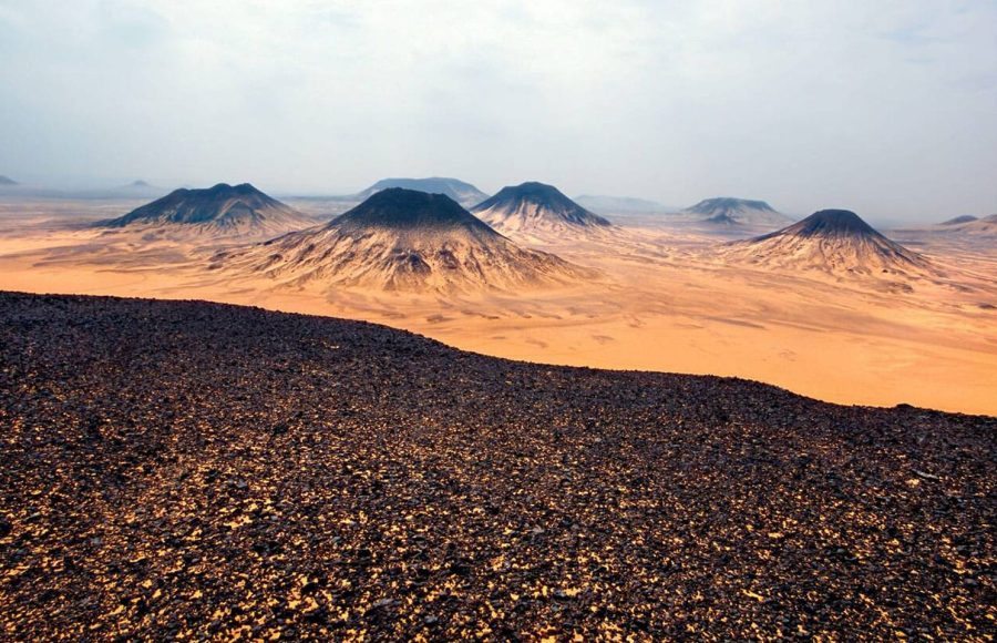 An expansive view of the Black Desert in Egypt, showing numerous conical, volcano-shaped black mounds contrasting with the ochre-colored desert sand under an overcast sky.