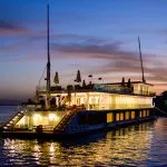A luxury two-deck Dahabiya boat illuminated with warm lights on the Nile River at sunset.