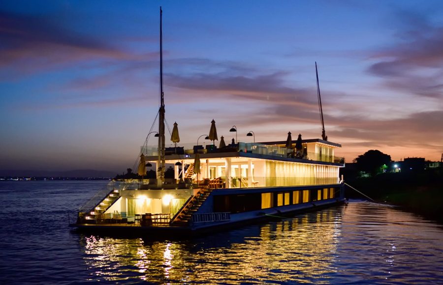 A luxury two-deck Dahabiya boat illuminated with warm lights on the Nile River at sunset.